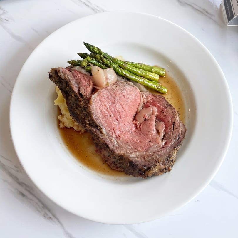 Prime rib with mashed potatoes, asparagus, and au jus on a white plate.