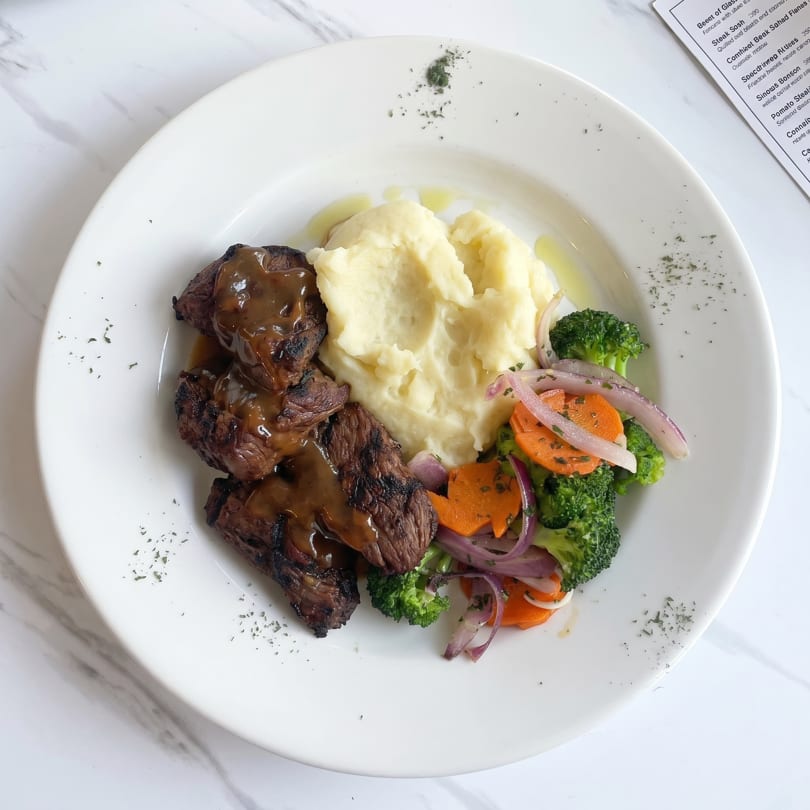 Grilled steak with gravy, mashed potatoes, and a side of broccoli, carrots, and red onions.