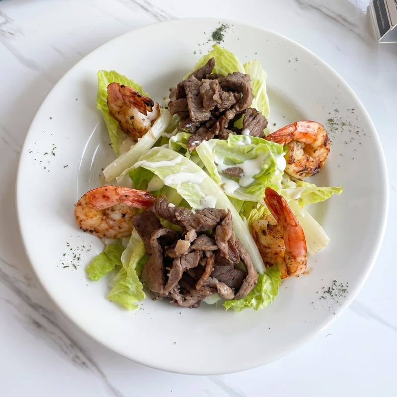 A white plate holds a salad with lettuce, grilled shrimp, and sliced beef, drizzled with dressing.